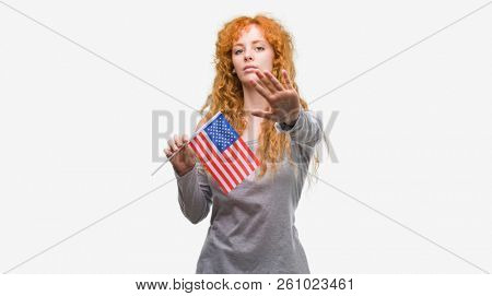 Young redhead woman holding flag of United States of America with open hand doing stop sign with serious and confident expression, defense gesture