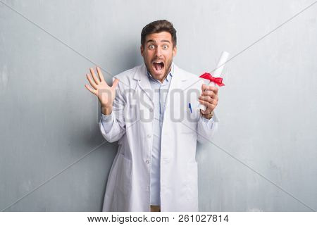 Handsome young doctor man over grey grunge wall holding diploma very happy and excited, winner expression celebrating victory screaming with big smile and raised hands