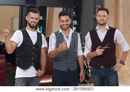Three Handsome Barbers Posing And Smiling. Bearded Hairdressers Standing In Barbershop And Holding S