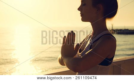 Young Woman Practicing Yoga Outdoors By The Sea At Sunset. Girl Standing With Eyes Closed And Prayer