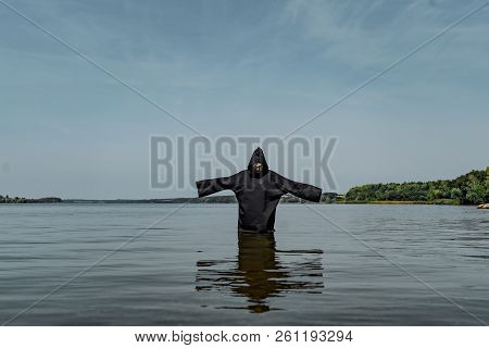 A Man In A Black Robe With Hands Outstretched Stands In The Middle Of The Lake In Warm Weather. His 