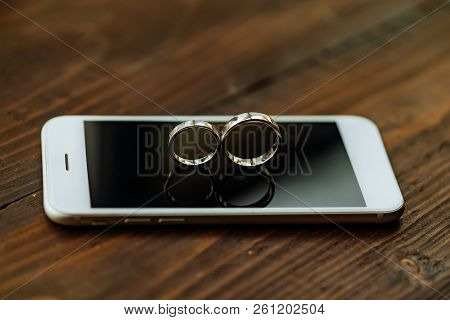 The Phone Is On A Wooden Table. Wedding Rings In The Form Of Eight Is On The Screen Of The Phone In 