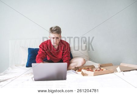 Young Attractive Man Sitting On A Bed In A Bedroom With A Pizza Box And Using A Laptop. Teenager Use