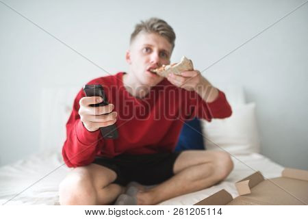 Young Man In A Red Sweatshirt Sits At Home On The Bed, Bites A Piece Of Pizza And Switches Channels 