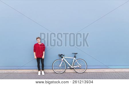 Stylish Young Man In A Red Sweatshirt Standing Beside A White Bicycle On A Blue Background And Looki