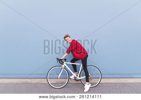 Happy Young Man In A Red Sweatshirt Sitting On A White Bike Against A Blue Wall Background. Stylish 