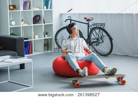 Young Asian Man In Headphones Sitting In Bean Bag Chair And Using Smartphone At Home