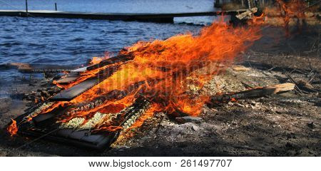 Fire Burning In The Wind By The Lake Near Blue Water. Wooden Boat Dock And Bridge Visible On The Bac