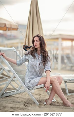 Romantic young beautiful girl with long fair hair in short dress sitting in chaise-longue on the beach. Attractive sleepy calm woman dreaming at dawn on the shore in Italy, Europe. Soft focus