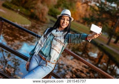 Modern Cute Girl Enjoying To Take A Selfie On The Bridge In Autumn City Park.