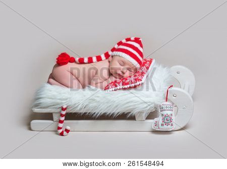 Charming infant baby in striped bonnet sleeping on red pillow on small wooden sledge crib with plush Christmas cane toy nerby
