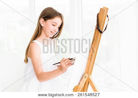 Pretty girl with brush in hand. Creative teen girl paitning a picture on easel. Girl practising drawing in art school