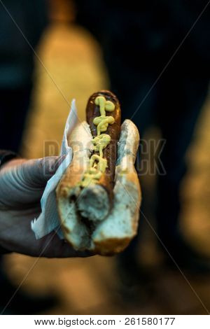 Person Hand Holding Bratwurst Or Rostbratwurst A Traditional German Sausage, With Mustard On A Bun O