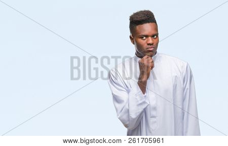 Young arabic african man wearing traditional djellaba over isolated background with hand on chin thinking about question, pensive expression. Smiling with thoughtful face. Doubt concept.