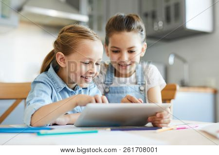 Two little girls having fun while watching curious vids or cartoons in touchpad in the kitchen