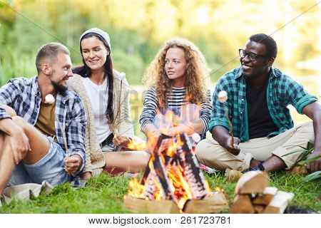 Group of young campers sitting by campfire and frying marshmellows while talking to each other