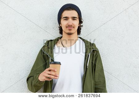 Waist Up Shot Of Stylish Teenager With Culry Dark Hair, Wears Fashionable Hat And Green Jacket, Hold