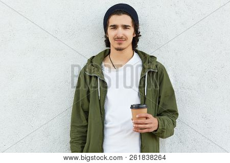Waist Up Shot Of Bearded Cool Curly Boy In Black Hat, Green Jacket, Dtinks Coffee Or Cappuccino, Pos