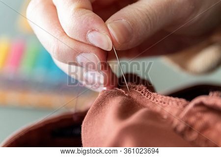 A Close Up Portrait Of A Hand Of Someone Holding A Needle, Sewing A Hole In A Pair Of Pants At The S