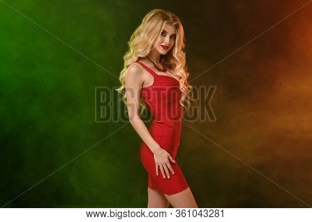 Curly Blonde Girl With Alluring Decollete, In Red Fitting Dress And Necklace. She Is Smiling, Posing