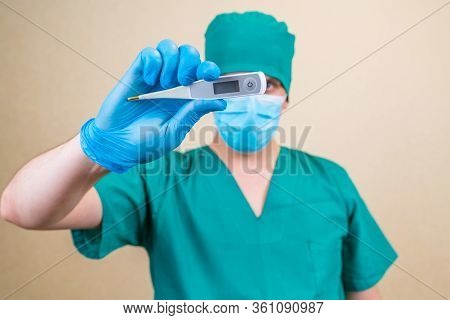 Doctor In Green Suit And Blue Mask Gloves With Thermometer In Hand On Yellow Background. Selective F