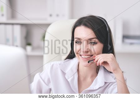 Young cheerful female customer support representative with headset looking at computer screen while consulting clients online