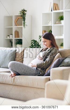 Happy young pregnant woman in casual outfit sitting with crossed legs on sofa in living room and talking to baby in her womb