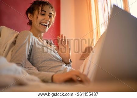 Young Woman Laughting And Talking On Video Call On Laptop Computer In Bed At Home, Sunlight In Bedro