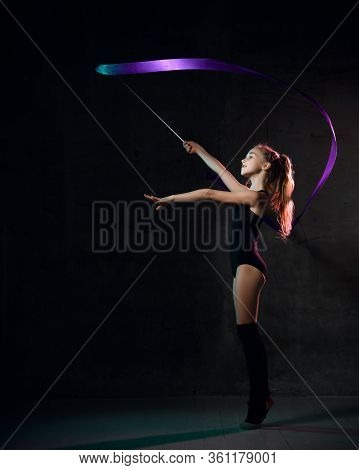 Young Girl Gymnast In Black Sport Body And Uppers Standing Sideways And Making Exercise With Blue Gy