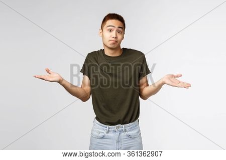 Who Knows, I Dont Aware. Portrait Of Confused Shrugging Asian Man In Greey T-shirt, Smirk And Apolog