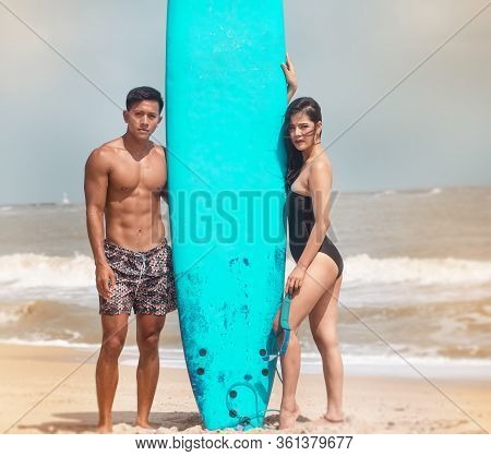 Young Handsome Man And Beautiful Woman Wearing Black Swimming Suit Posing Beside Serf Board On The B