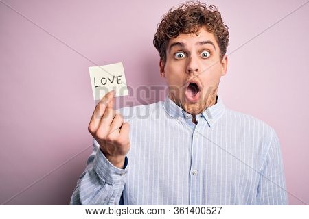 Young blond romantic man with curly hair holdiing paper with love message for valentines day scared in shock with a surprise face, afraid and excited with fear expression