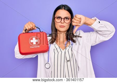 Young beautiful brunette doctor woman wearing stethoscope and coat holding first aid kit with angry face, negative sign showing dislike with thumbs down, rejection concept