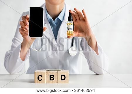 Cropped View Of Doctor Holding Smartphone With Blank Screen And Oil Bottle Near Cubes With Cbd Lette