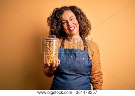 Middle age beautiful woman holding jar with macaroni pasta over isolated yellow background with a happy face standing and smiling with a confident smile showing teeth