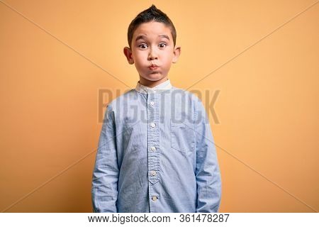 Young little boy kid wearing elegant shirt standing over yellow isolated background puffing cheeks with funny face. Mouth inflated with air, crazy expression.