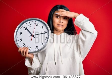Young beautiful chinese woman holding big clock standing over isolated red background stressed with hand on head, shocked with shame and surprise face, angry and frustrated. Fear and upset for mistake