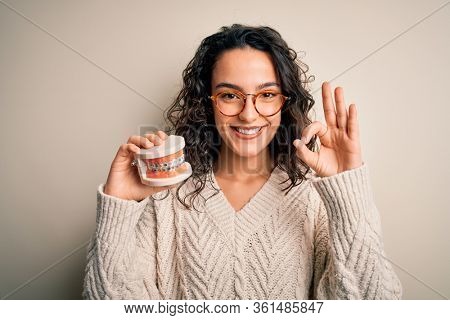 Young beautiful woman with curly hair holding plastic teeth with dental baces doing ok sign with fingers, excellent symbol