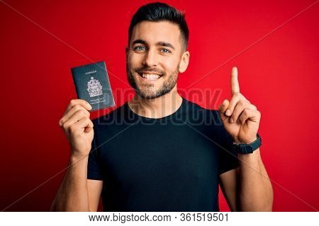 Young handsome tourist man holding canada canadian passport id over red background surprised with an idea or question pointing finger with happy face, number one