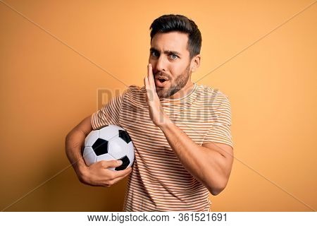 Handsome player man with beard playing soccer holding footballl ball over yellow background hand on mouth telling secret rumor, whispering malicious talk conversation
