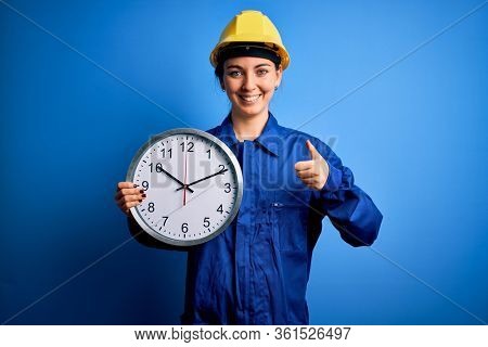 Beautiful blonde worker woman with blue eyes wearing security helmet holding big clock happy with big smile doing ok sign, thumb up with fingers, excellent sign