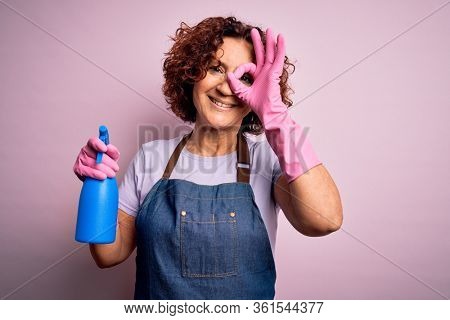 Middle age curly hair woman cleaning doing housework wearing apron and gloves using spayer with happy face smiling doing ok sign with hand on eye looking through fingers