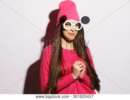 fashion portrait of trendy casual young woman in pink pulover and hat, big sunglasses, posing over grey background.