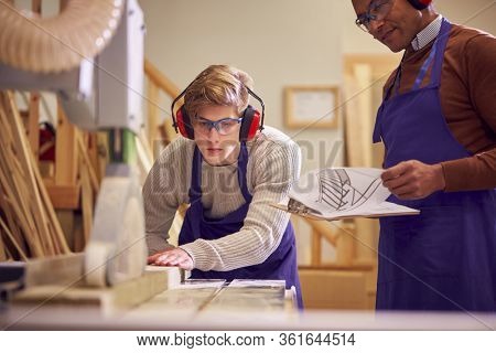 Tutor With Male Carpentry Student In Workshop Studying For Apprenticeship At College Using Bench Saw