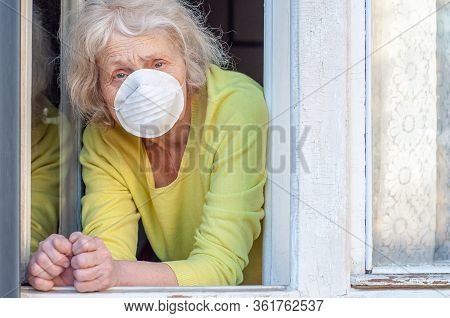 A Sad Elderly Wrinkled Woman Breathes Fresh Air Through The Window In The Respiratory Mask Of Her Ho