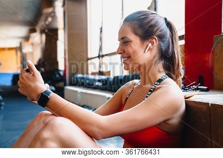 Portrait of cheerful cute sportswoman using wireless earphone and cellphone while sitting in athletic hall