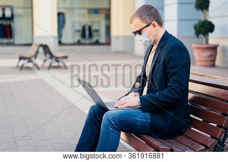 Sideways Shot Of Busy Man Sits On Wooden Bench Outdoor At Station, Waits For Transport, Uses Modern 