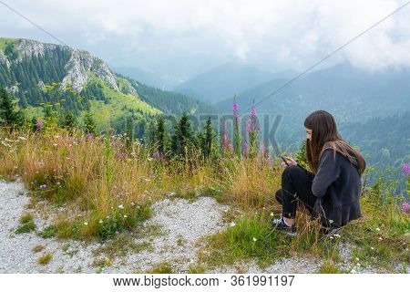 Young Teenage Girl Looking At Selfphone On Mountain Top Among Pink Mountain Flowers Against Green An