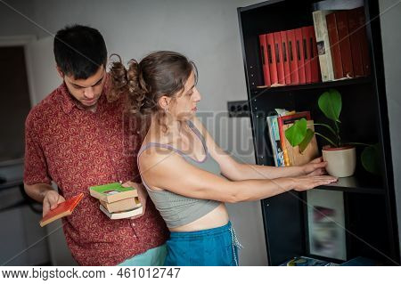 A Relaxed Young Couple Puts Various Kinds Of Book On A Shelf In The Room Of Their Apartment