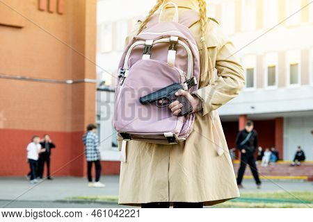 Firearms At School. Schoolgirl With A Gun In A Backpack On A School Background. The Girl Carries A G